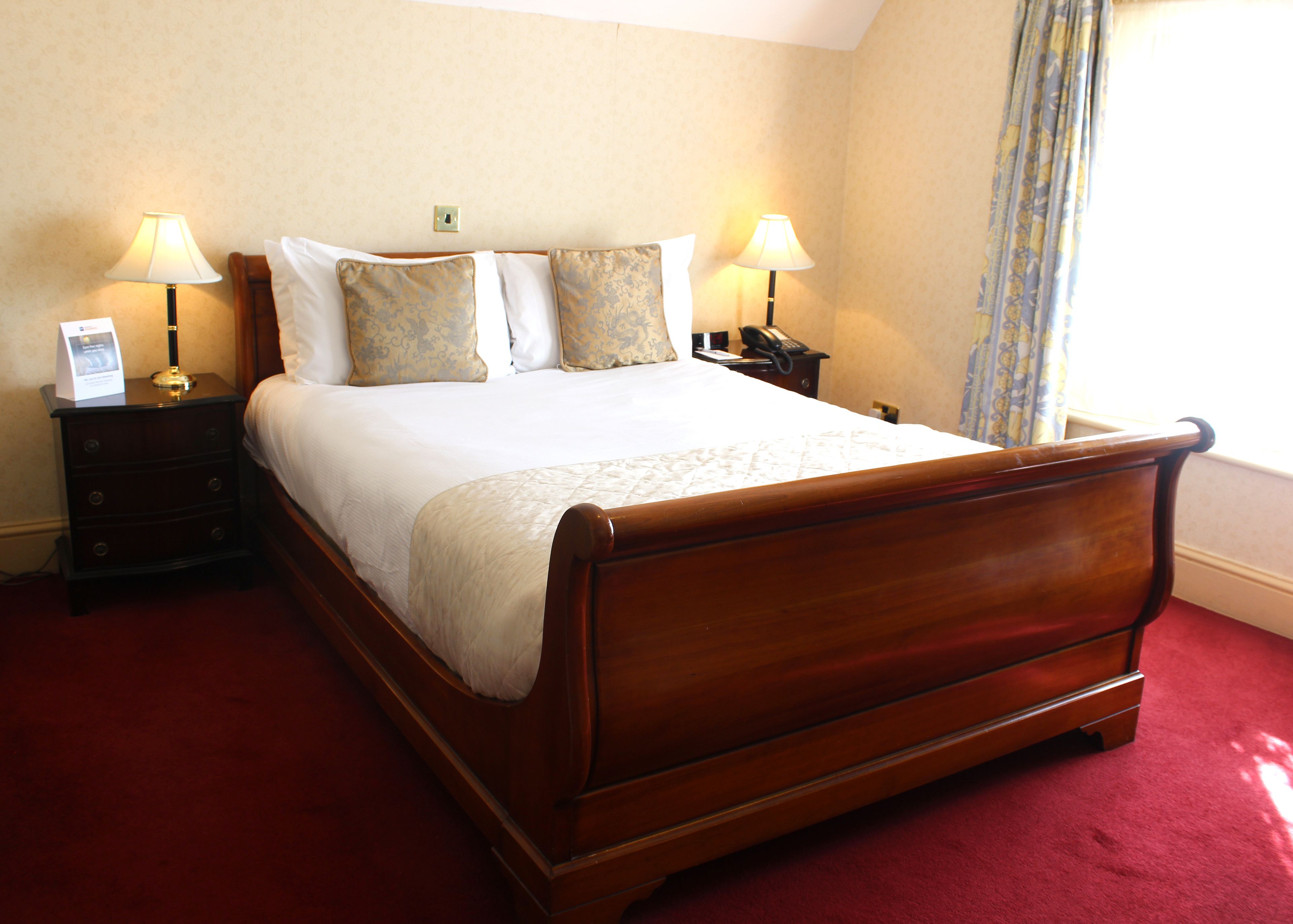A neatly made bed with a wooden frame, two bedside tables with lamps, and a red carpeted floor.