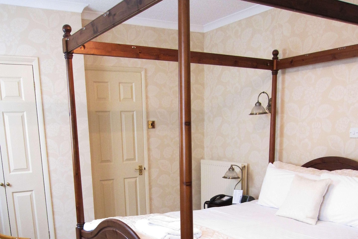 A four-poster bed with a quilted bedspread, a bedside table with a lamp, and a chair with a plaid cushion.