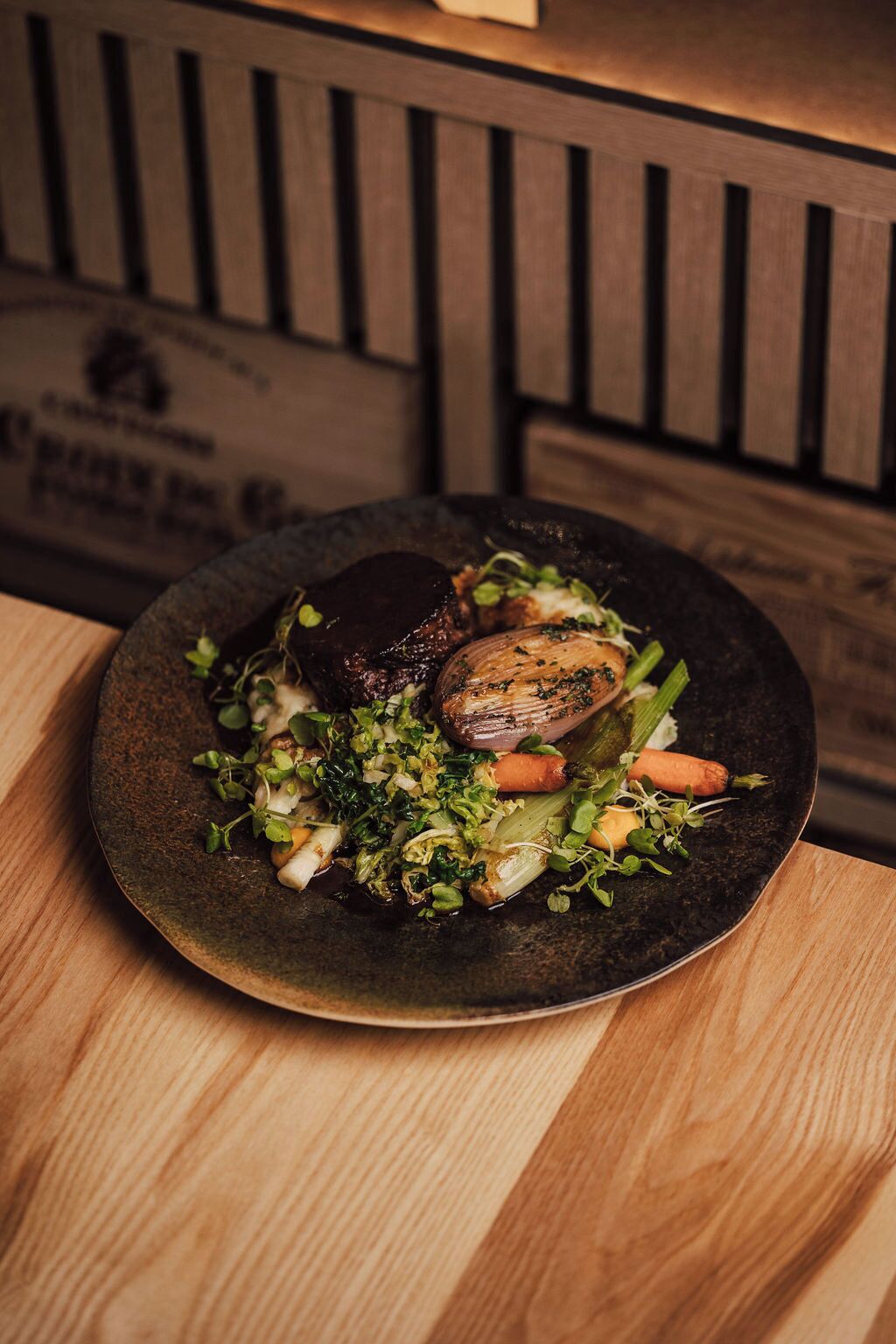 A plate of food with a variety of vegetables and a piece of meat on a dark plate, placed on a wooden table.