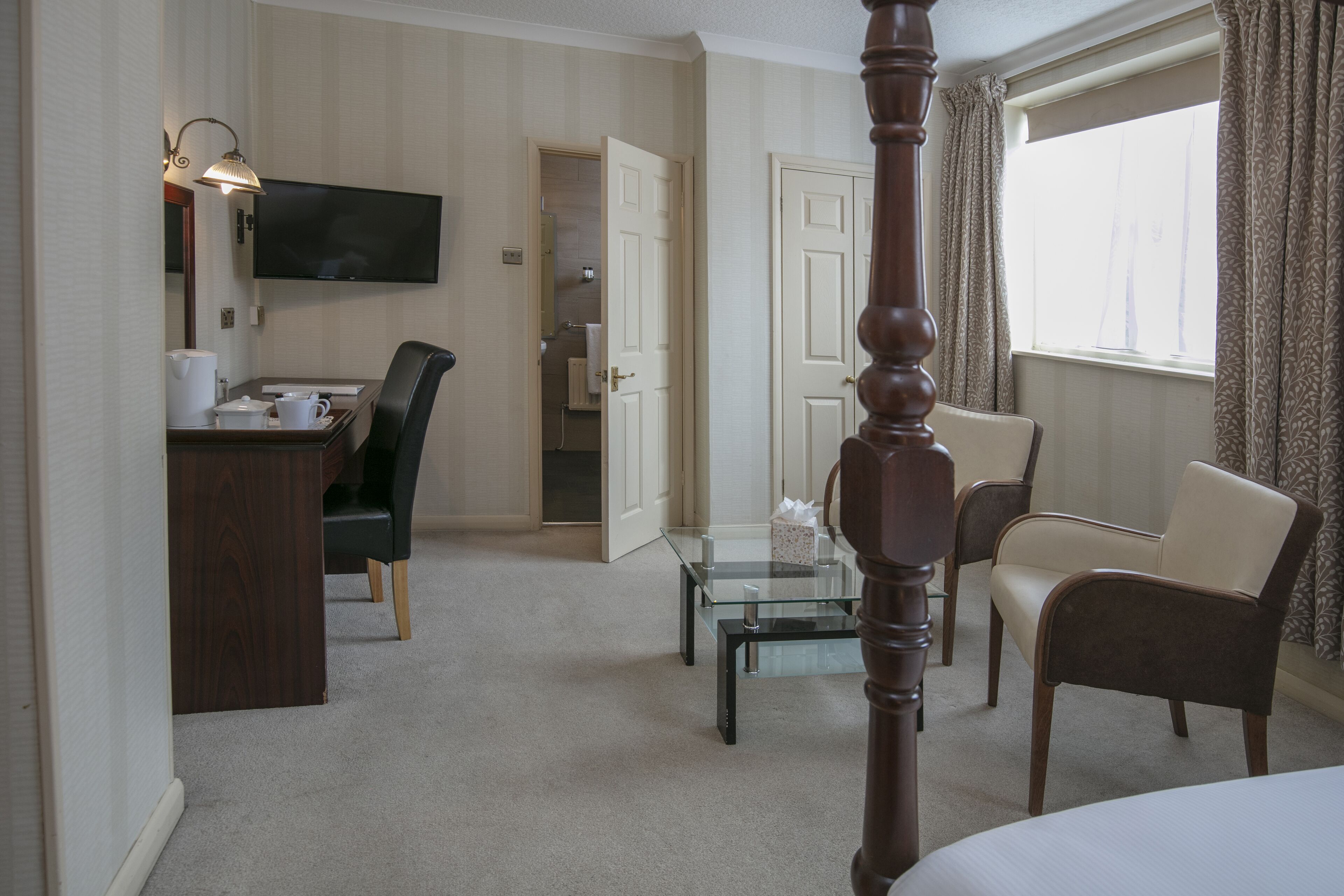 A hotel room with a bed, a desk with a chair, a glass coffee table, a TV, and a bathroom visible through an open door.