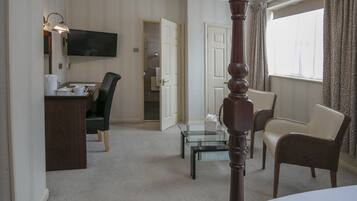 A hotel room with a bed, a desk with a chair, a glass coffee table, a TV, and a bathroom visible through an open door.