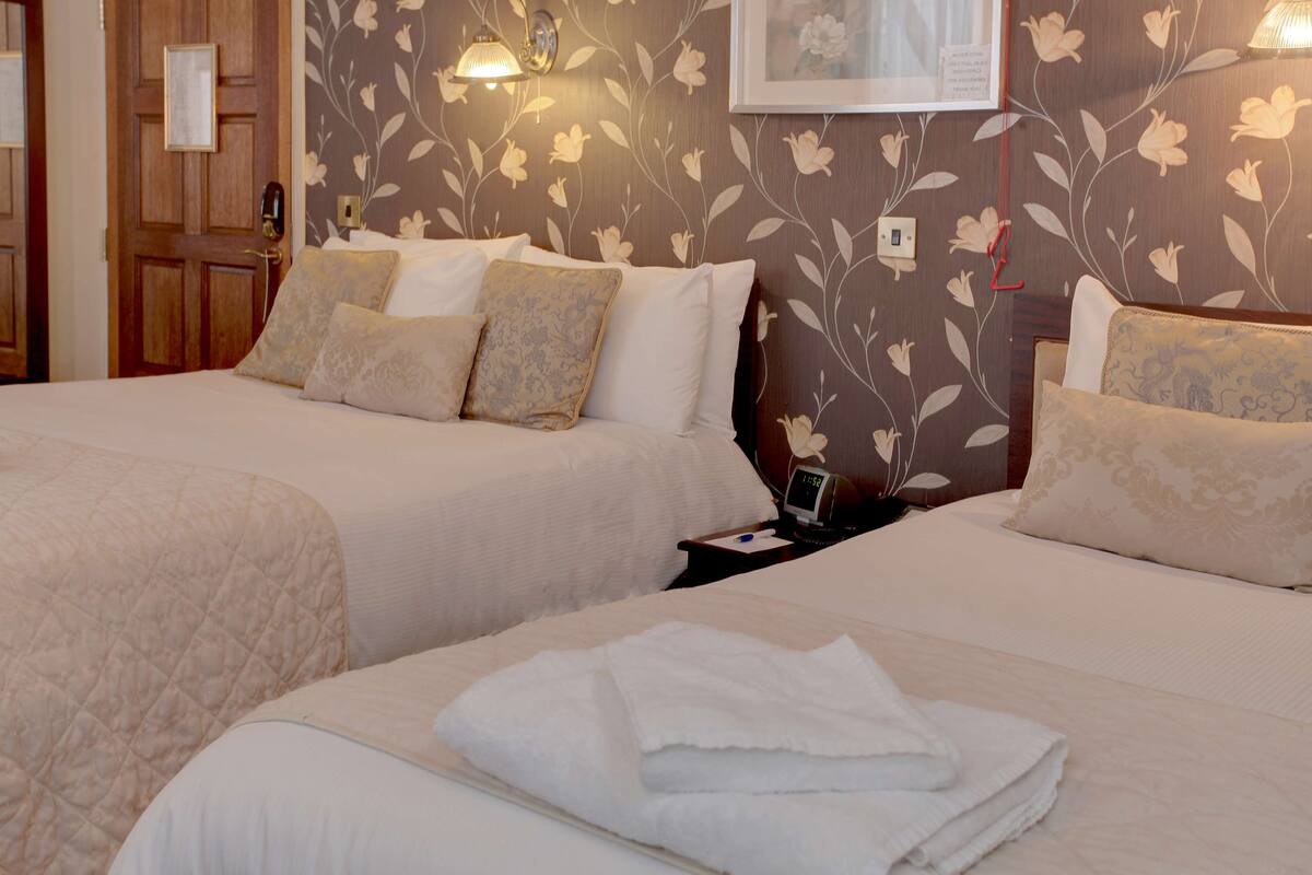 Une chambre d’hôtel avec deux lits, un papier peint à motifs floraux, une horloge et une table de chevet avec une lampe.