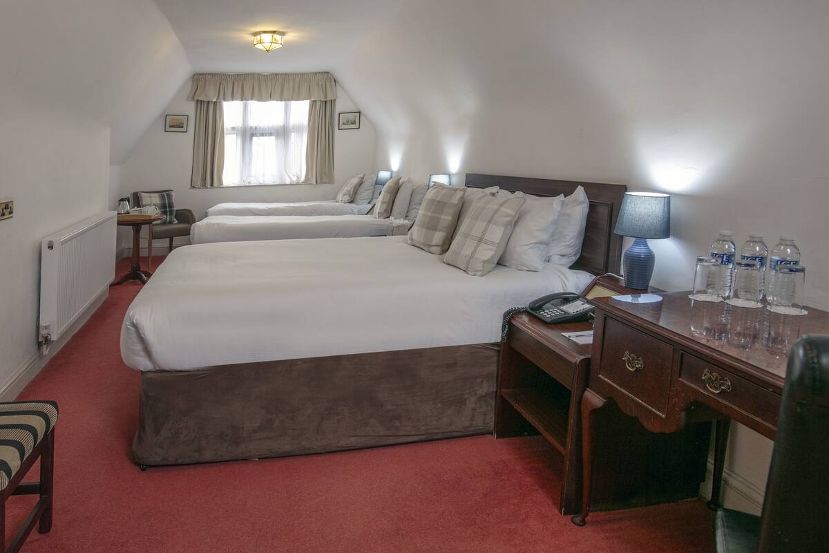 Une chambre d’hôtel avec trois lits simples, un bureau en bois, une chaise, un radiateur, une fenêtre avec des rideaux et une lampe de chevet.