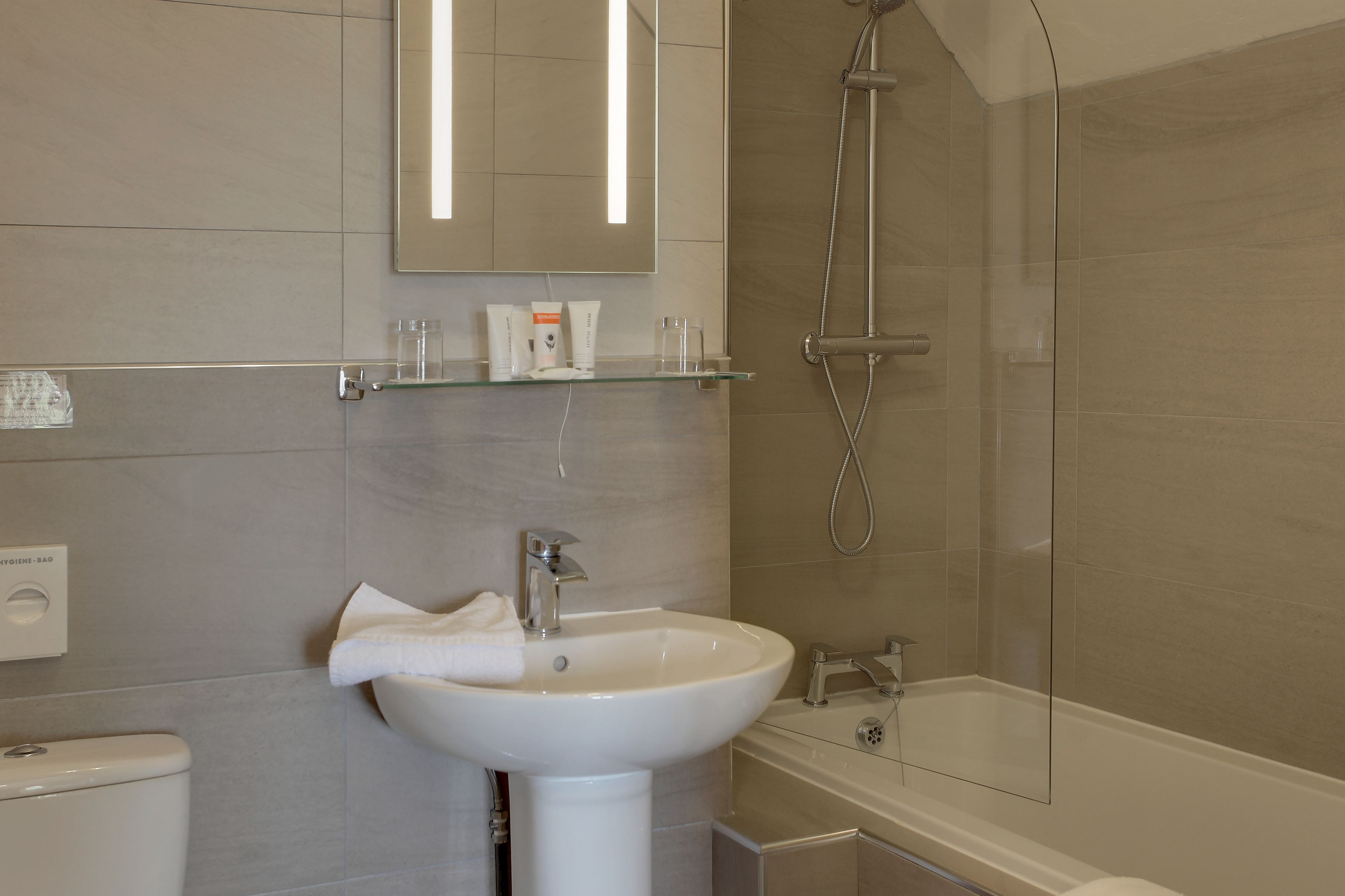 Un baño moderno con un lavamanos blanco, un estante de vidrio sobre el lavamanos, un inodoro y una bañera.