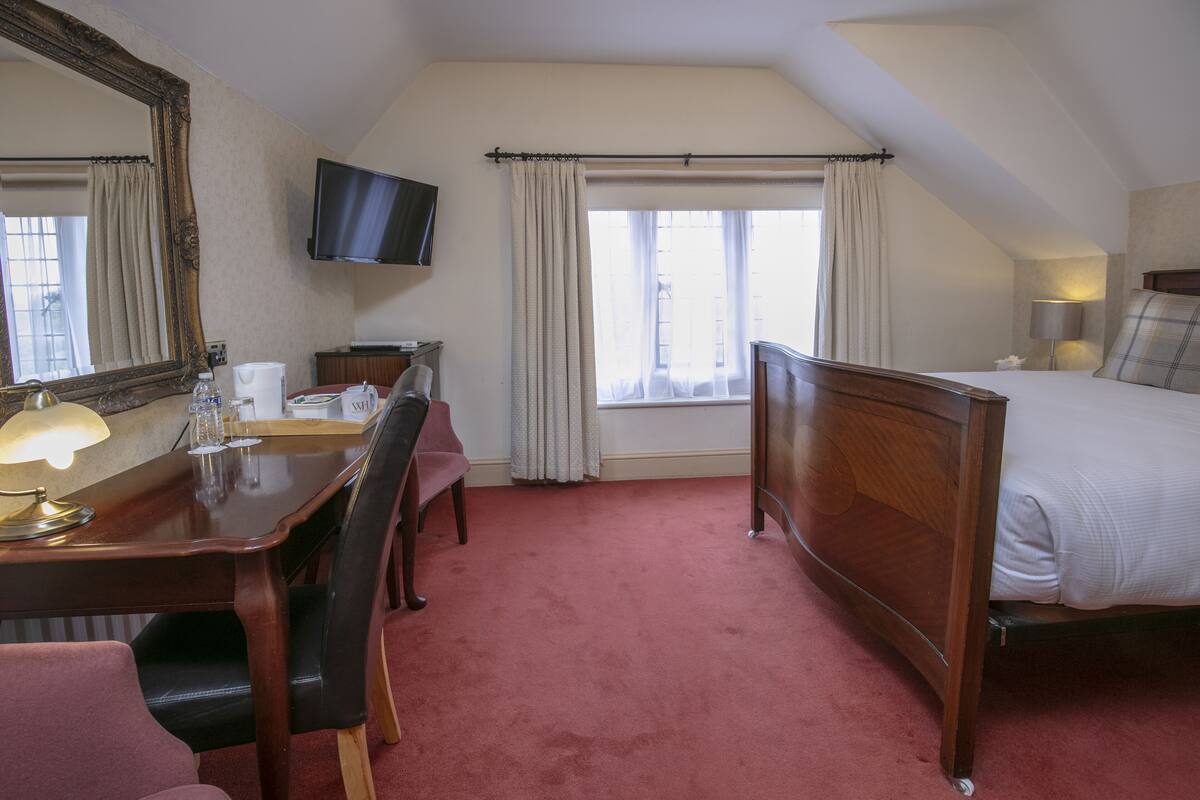 Une chambre d’hôtel avec un lit en bois, un bureau avec une lampe, une chaise, un téléviseur fixé au mur et une fenêtre avec des rideaux.