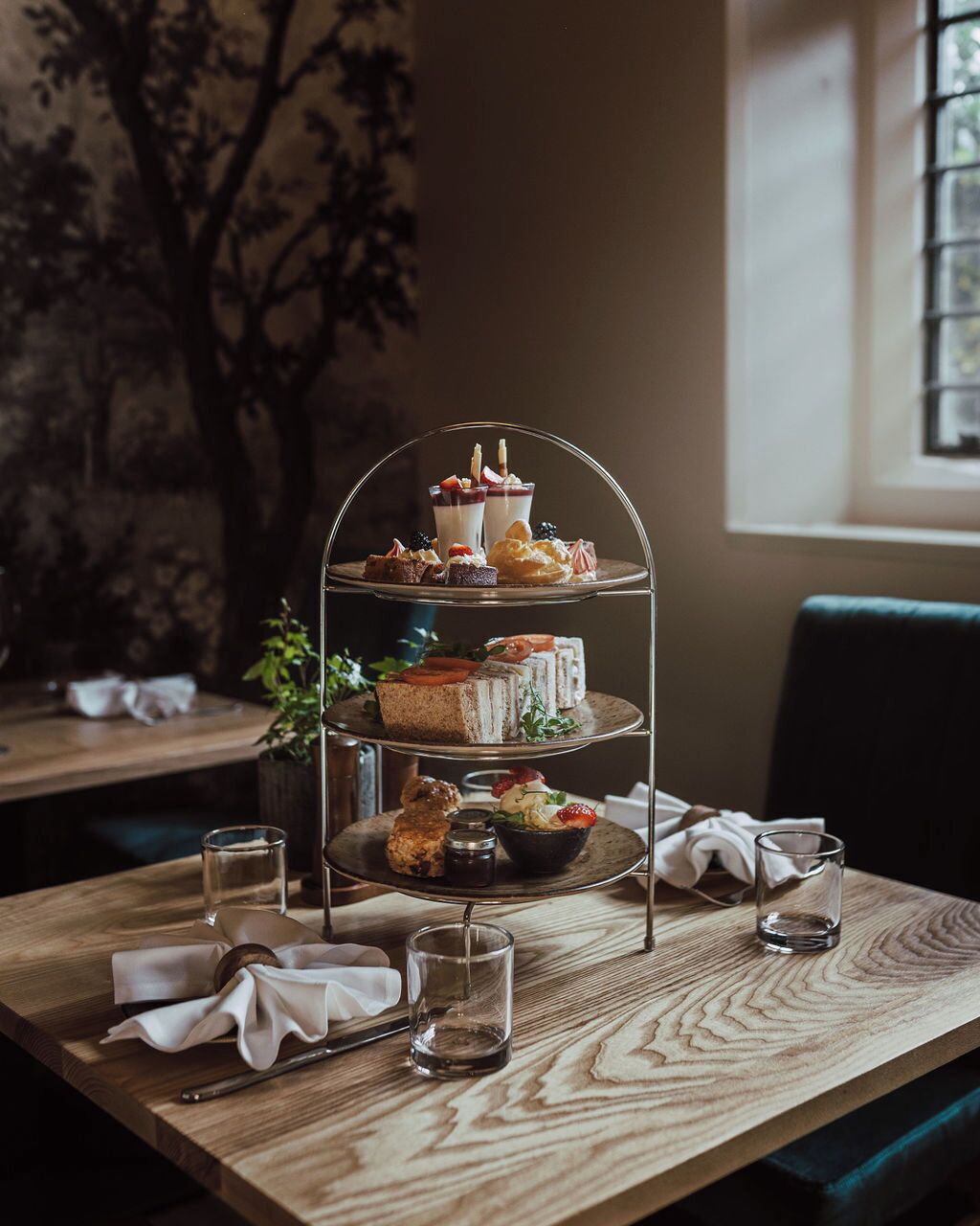 A three-tiered dessert stand with various pastries and drinks on a wooden table in a room with a window and a plant in the background.