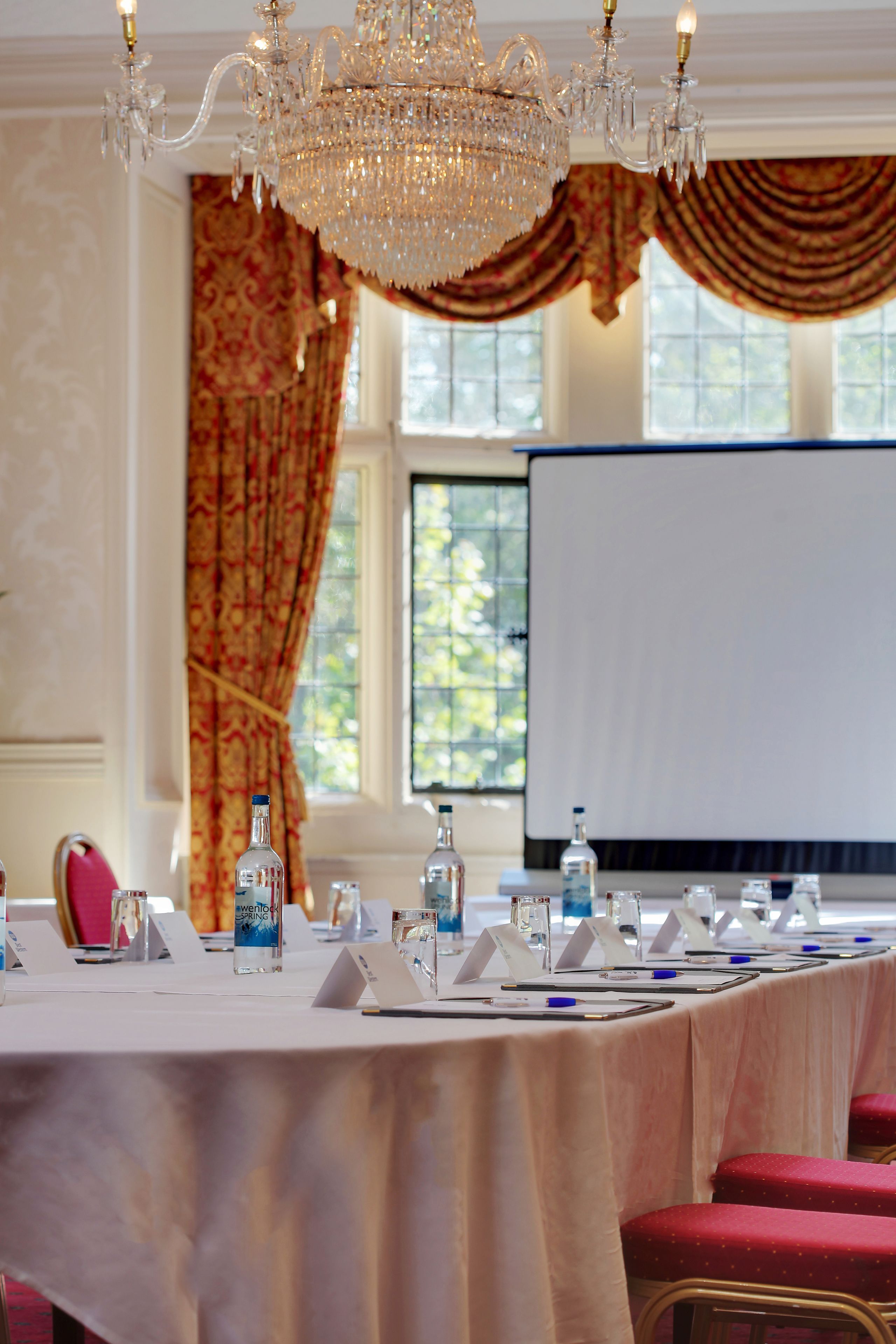 A conference room with a long table, chairs, a projector screen, and a chandelier.