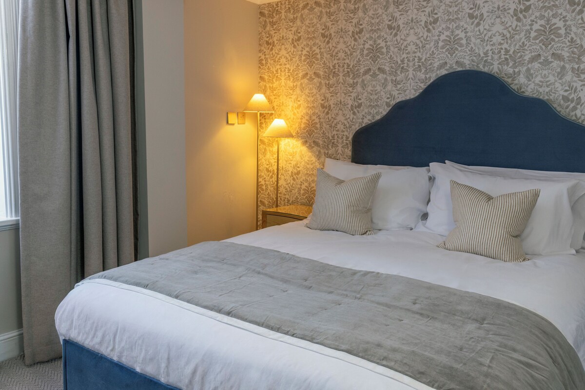 A neatly made bed with a blue headboard, two bedside lamps, and patterned wallpaper.