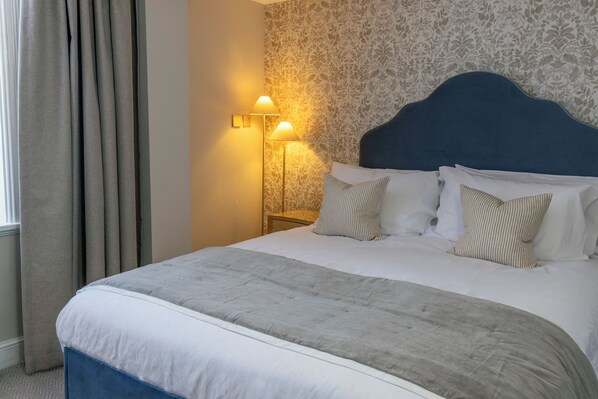 A neatly made bed with a blue headboard, two bedside lamps, and patterned wallpaper.