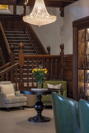 A staircase with wooden railings, a chandelier, a green sofa, a patterned armchair, and a table with a vase of flowers. - Newton Park County House Hotel (Burton on Trent)
