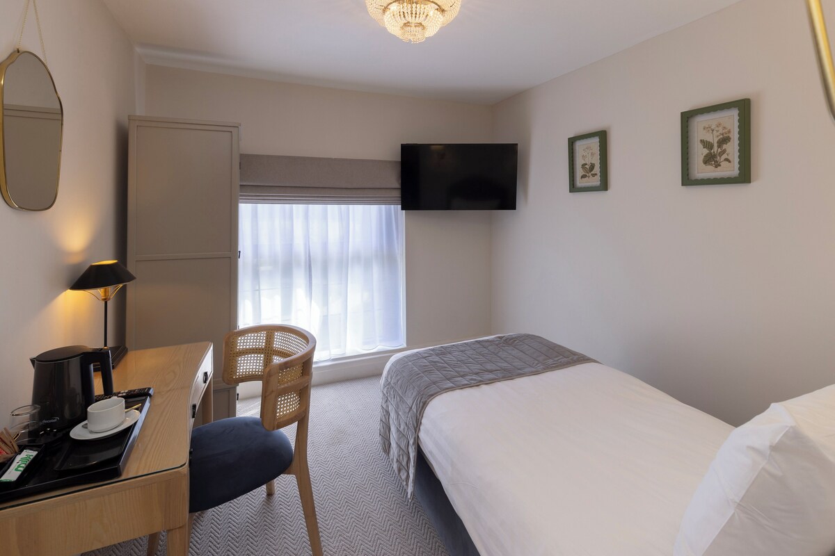 A hotel room with a bed, a desk with a coffee maker, a chair, a TV mounted on the wall, and two framed pictures.
