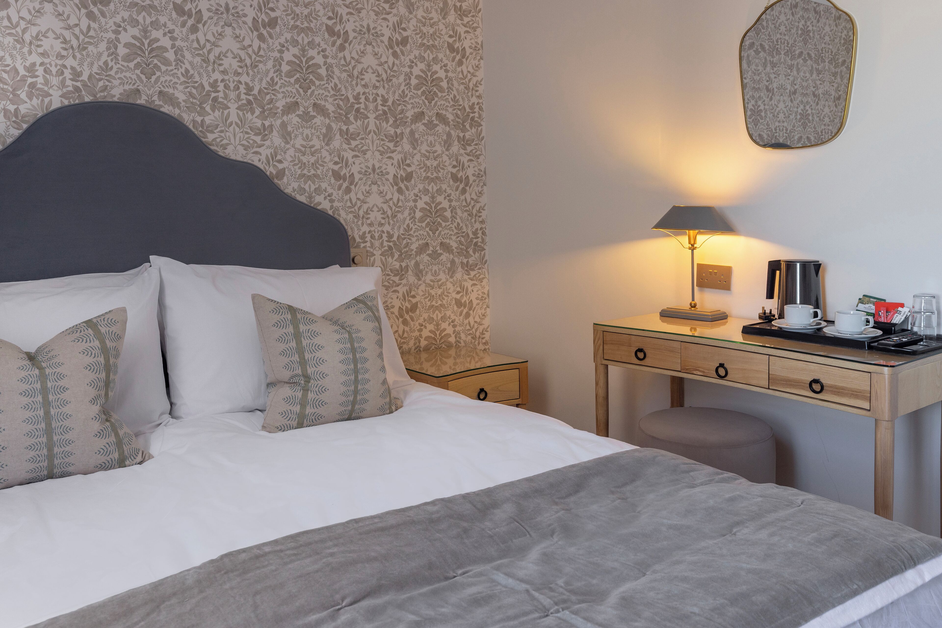 A neatly made bed with patterned pillows, a bedside table with a lamp, a coffee maker, and a small stool.