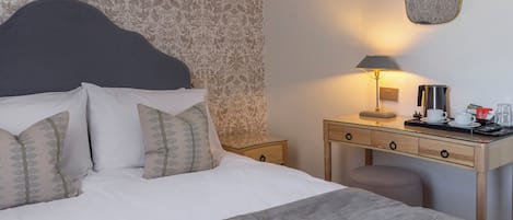 A neatly made bed with patterned pillows, a bedside table with a lamp, a coffee maker, and a small stool.