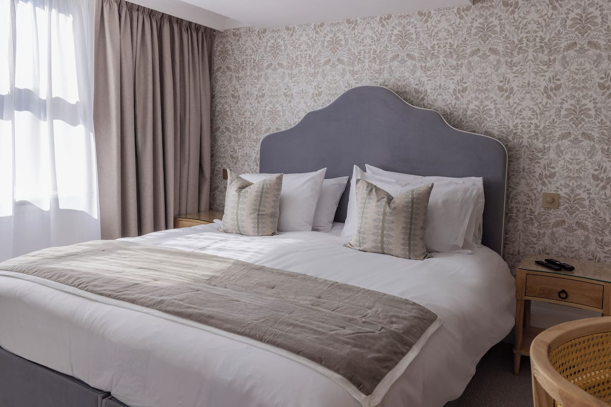 Una cama bien tendida con cabecera gris, ropa de cama blanca y almohadas estampadas. Se observa una mesita de noche con un control remoto y una silla de mimbre.