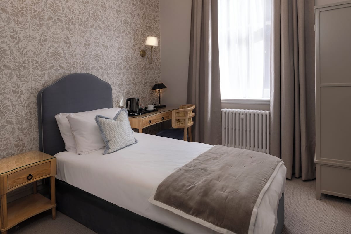 Una cama bien hecha con almohadas, una mesita de noche con una lámpara, una pequeña mesa con una cesta y un radiador.