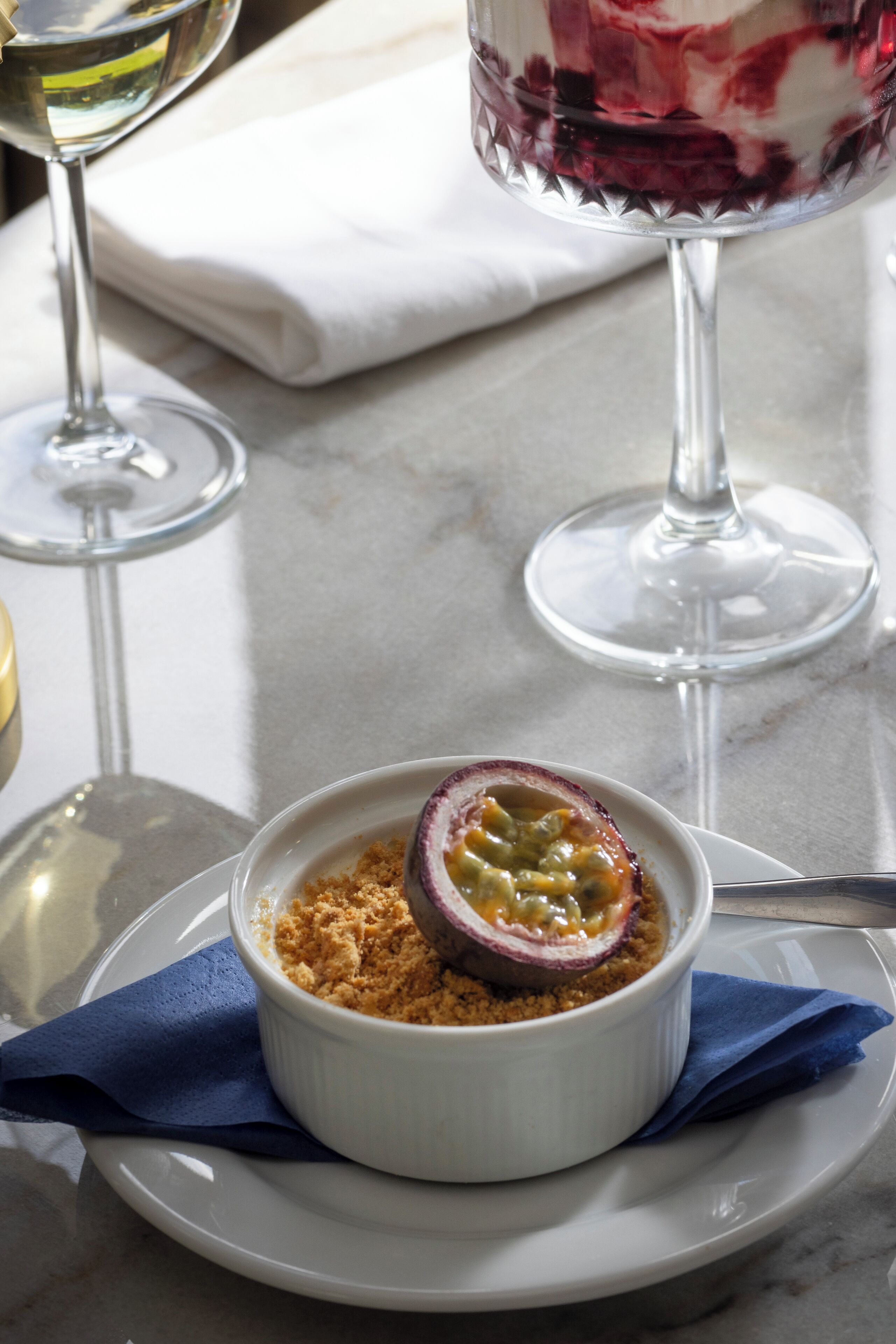 Un dessert servi dans un bol blanc, posé sur une assiette avec une serviette bleue, un verre de vin blanc et un verre de vin rouge, le tout sur une table en marbre.