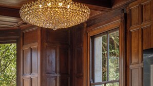 Una habitación con paneles de madera, una lámpara de araña grande, una cabeza de animal montada, muebles con diseños y vistas a través de un ventanal amplio.