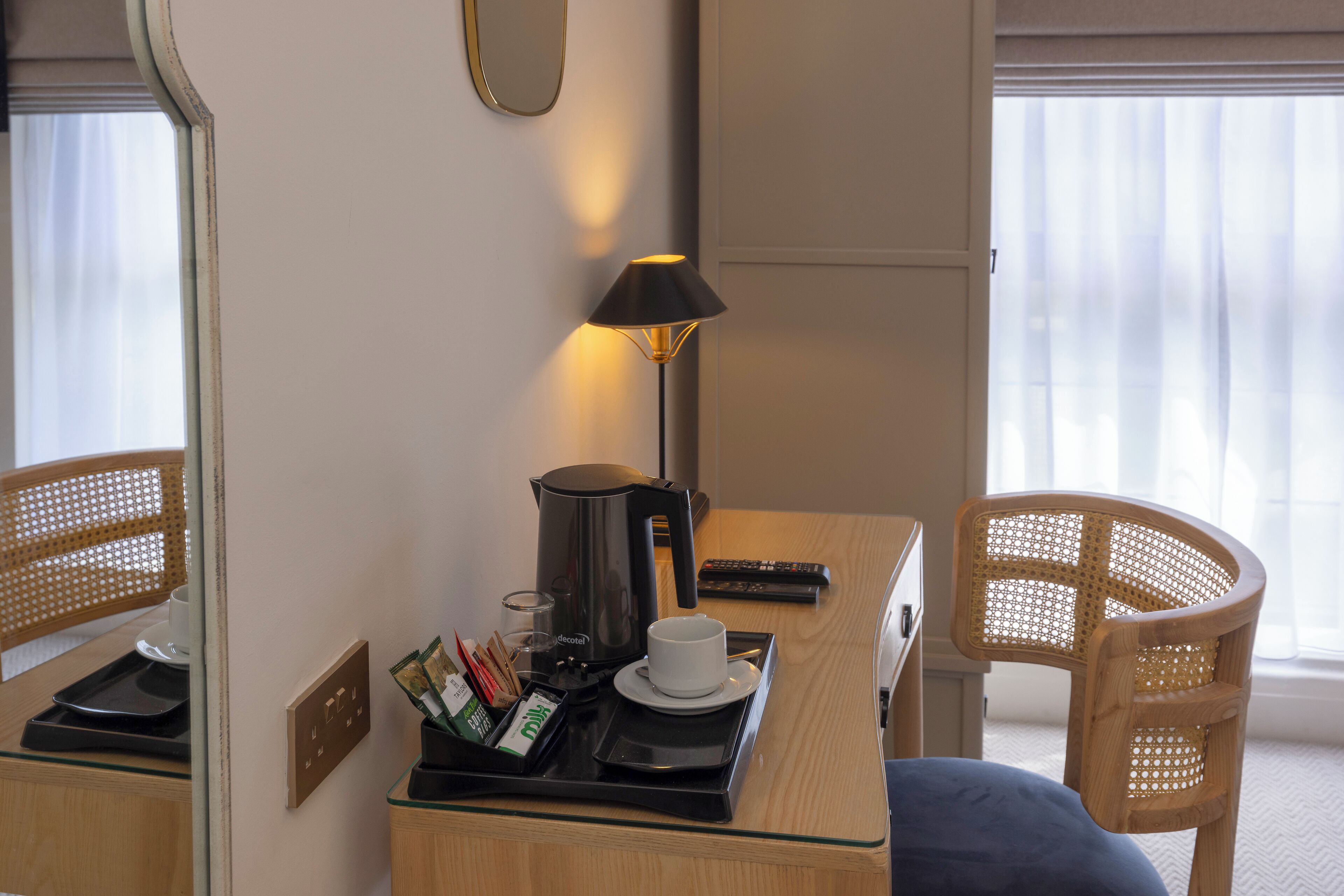 Une chambre d’hôtel équipée d’un bureau, d’une lampe, d’une machine à café, d’une tasse, d’un plateau avec divers articles, d’une télécommande et d’une chaise.