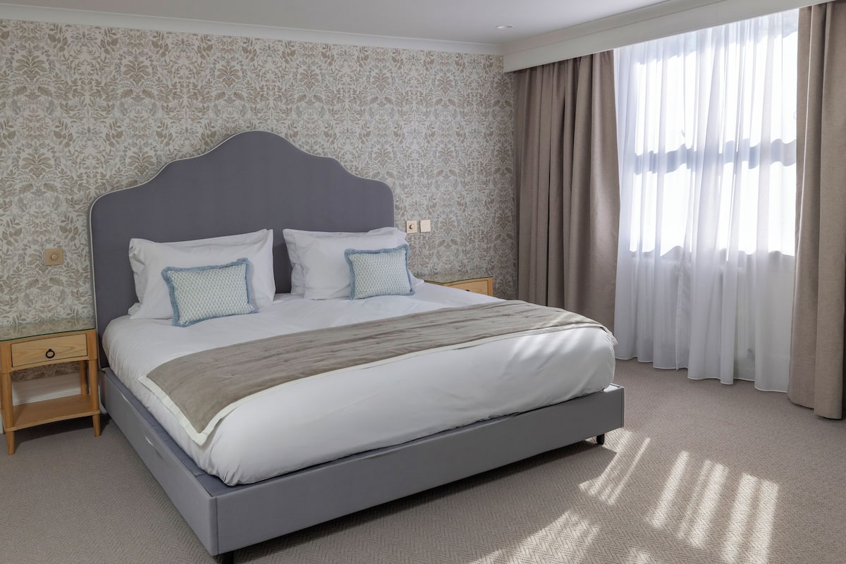 Habitación de hotel moderna con una cama grande, una mesita de noche, una ventana con cortinas y papel tapiz estampado.