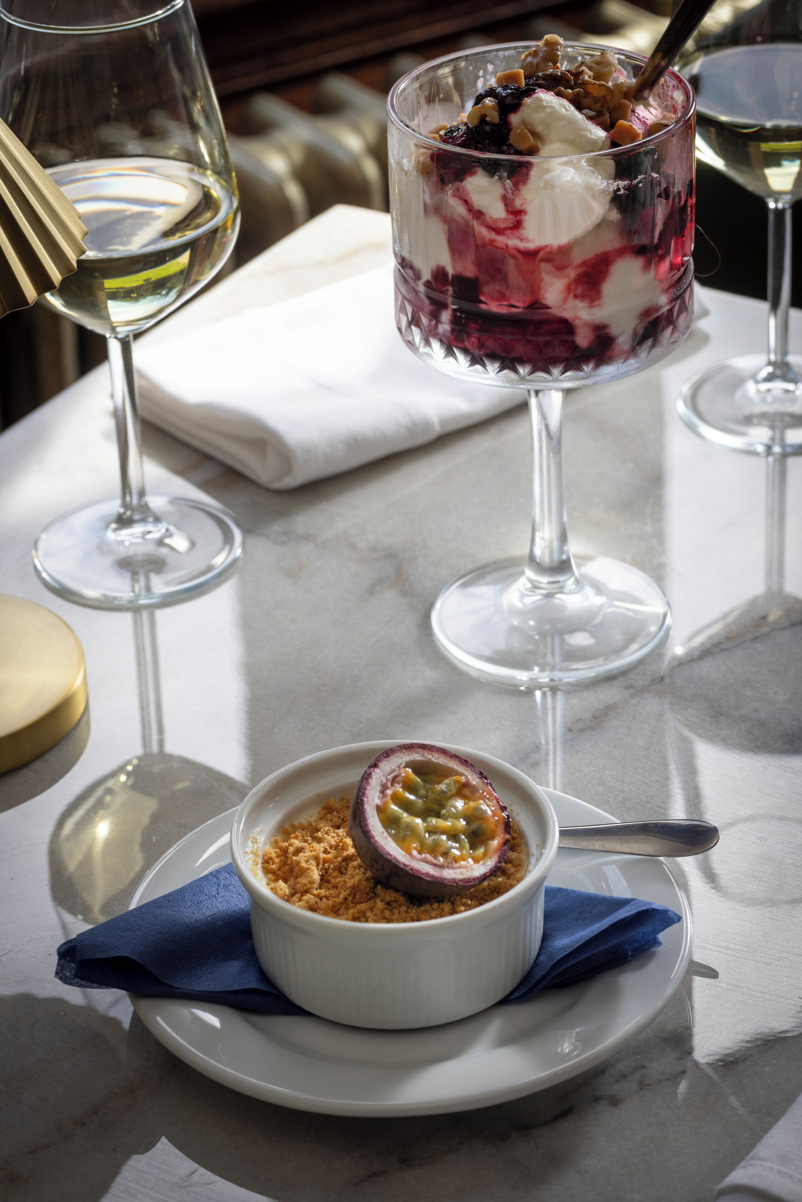 Un dessert composé d’une boule de glace et de fruits rouges, un saladier de fruits et un verre de vin posés sur une table en marbre.
