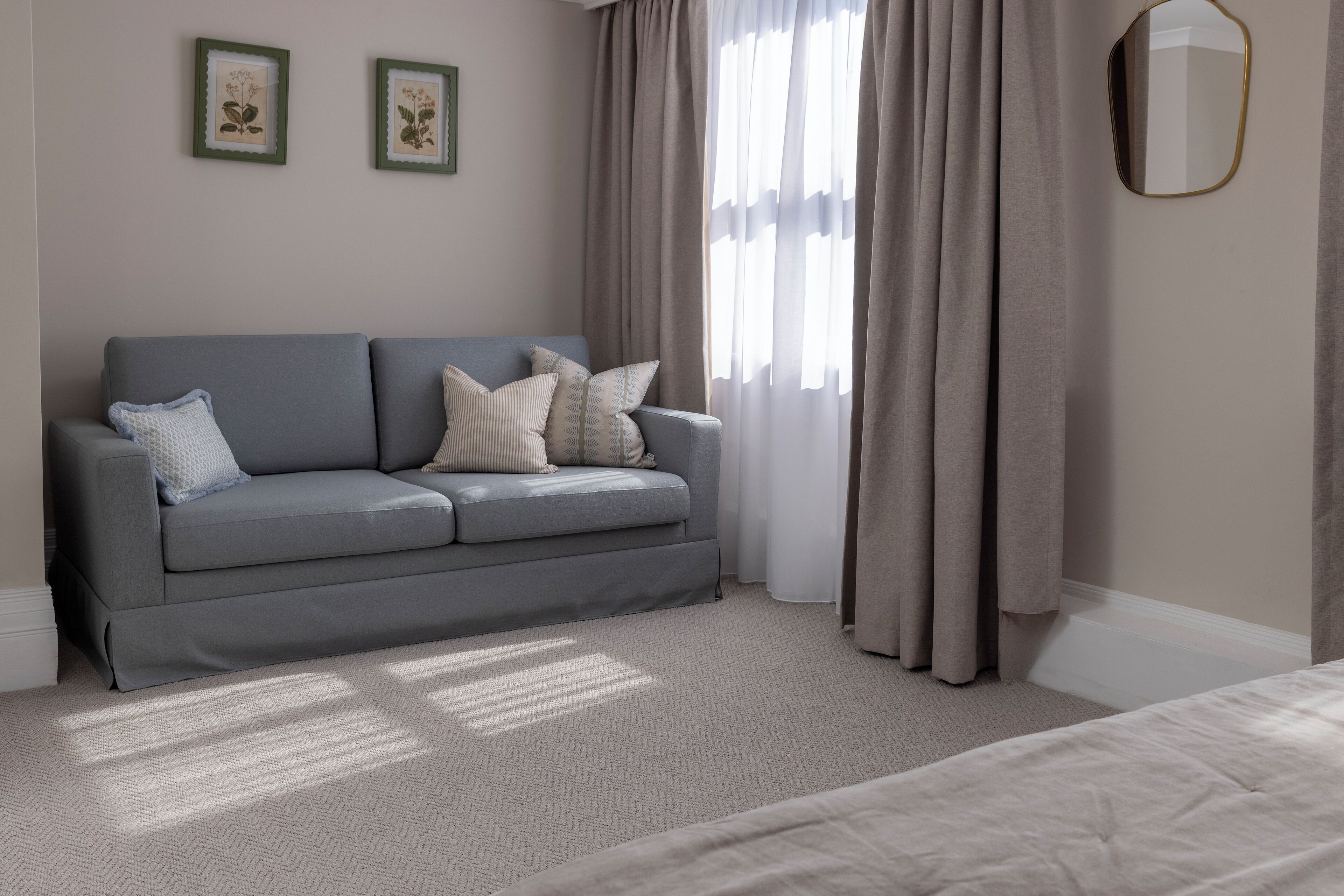 A neutral-colored room with a grey sofa, a bed, and a window with curtains.