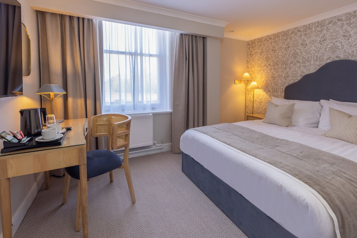 A hotel room with a bed, a desk, a chair, a window with curtains, and a wall with a floral pattern.