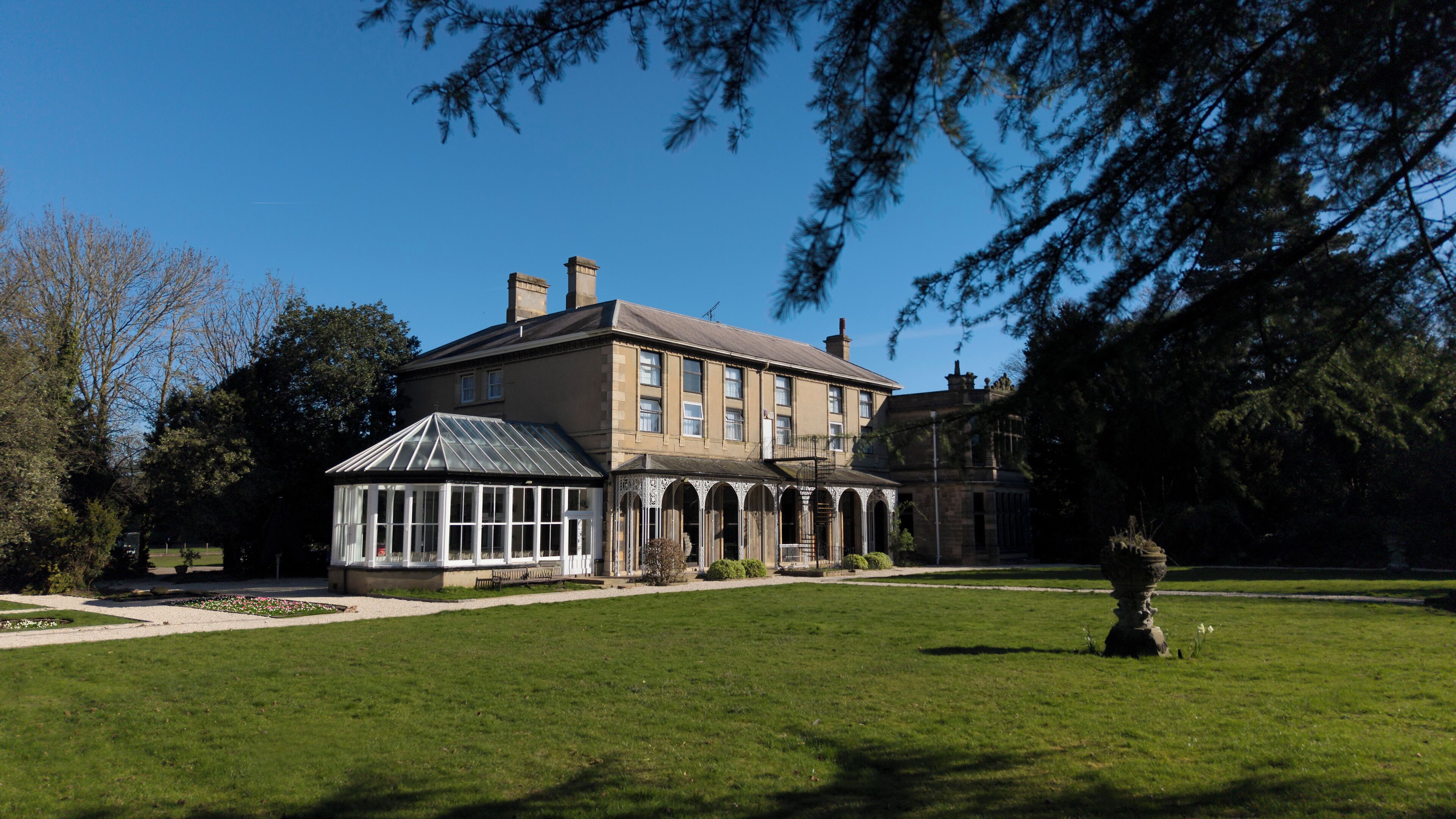 A large, historic building with a conservatory, surrounded by a well-maintained lawn and trees.