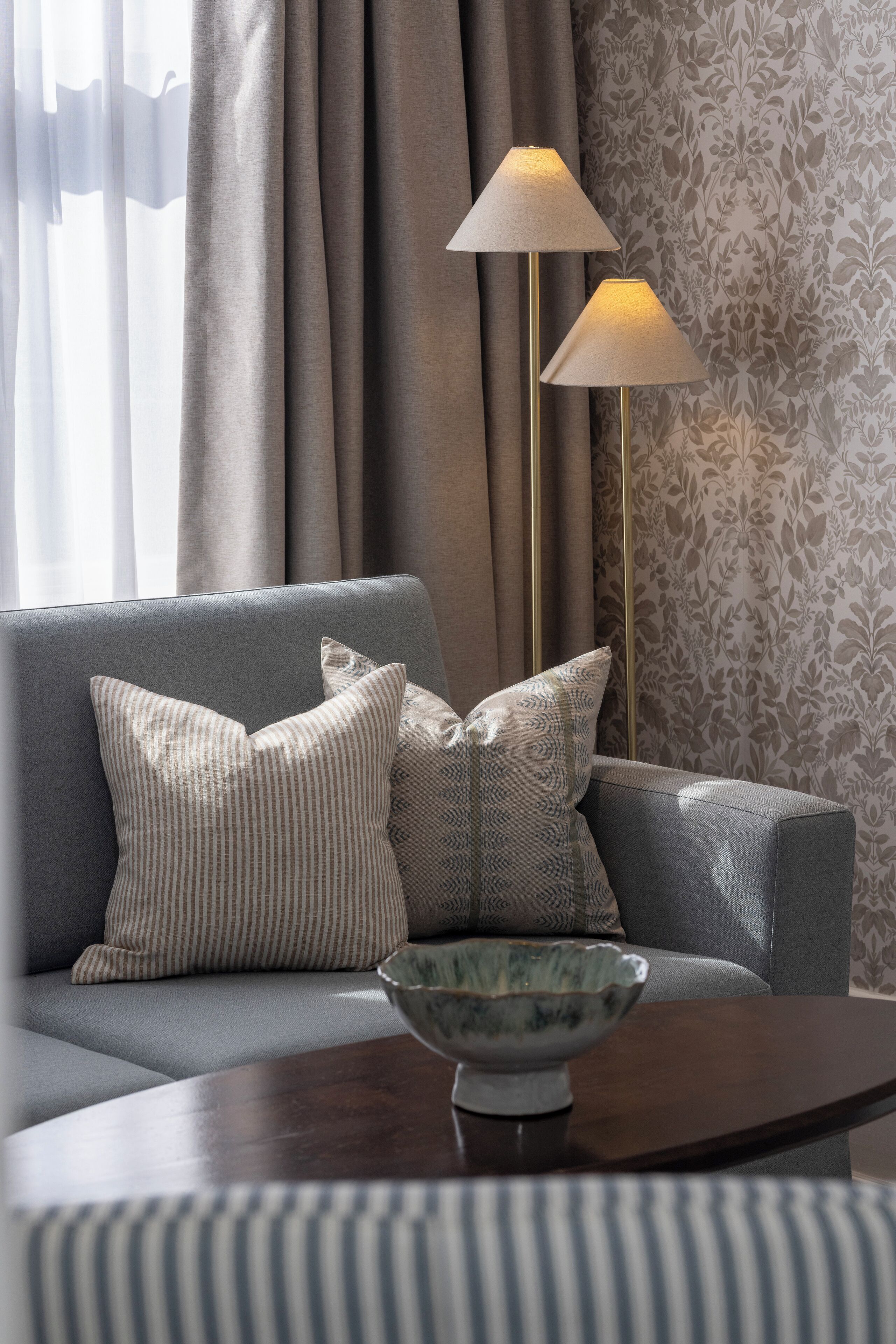 A modern living room with a grey sofa, striped pillows, a bowl on a wooden table, and a patterned wallpaper.