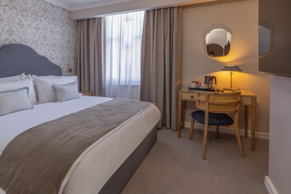 Quarto de hotel com uma cama grande, uma escrivaninha com abajur, uma cadeira e um espelho na parede.