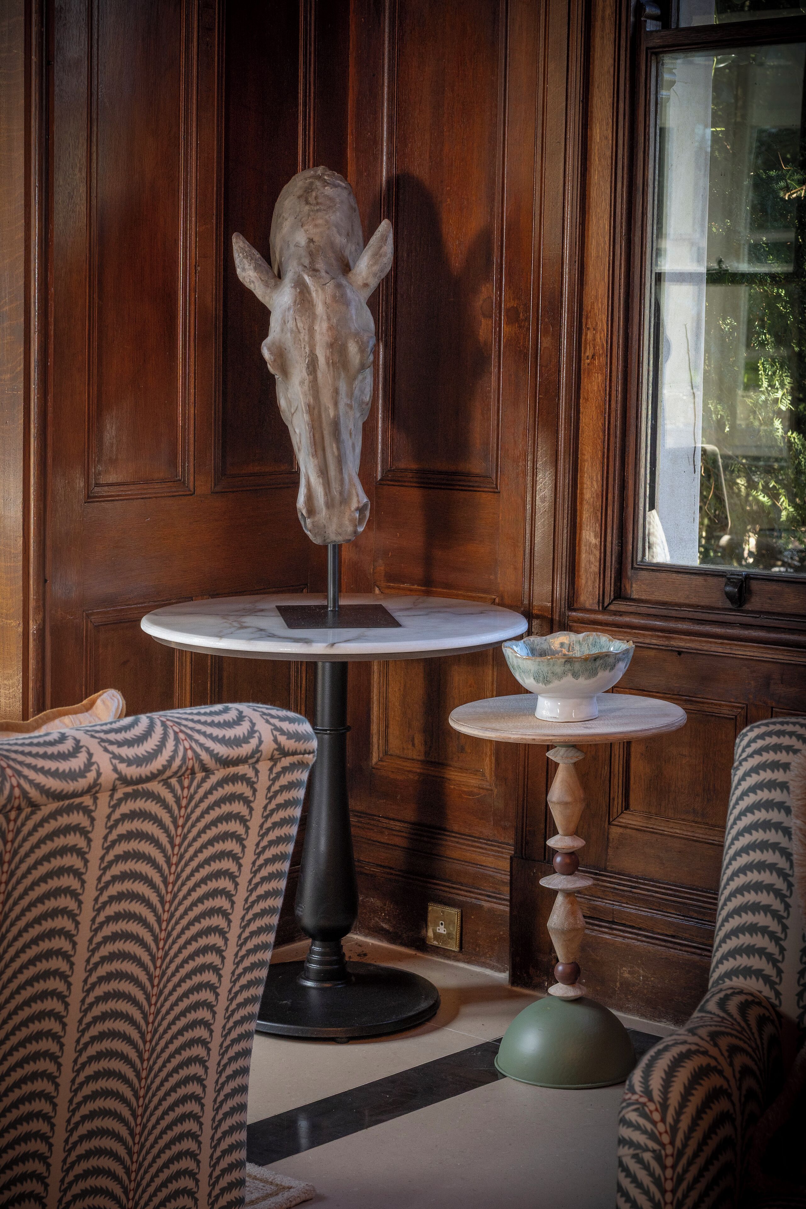 A room with a wooden paneled wall, a round table with a bowl, patterned chairs, and a head sculpture.