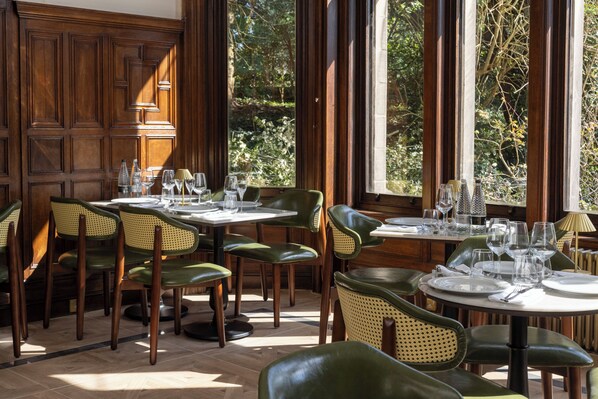 A dining area with wooden paneled walls, large windows with a view of trees, and green upholstered chairs. - Newton Park County House Hotel (Burton on Trent)
