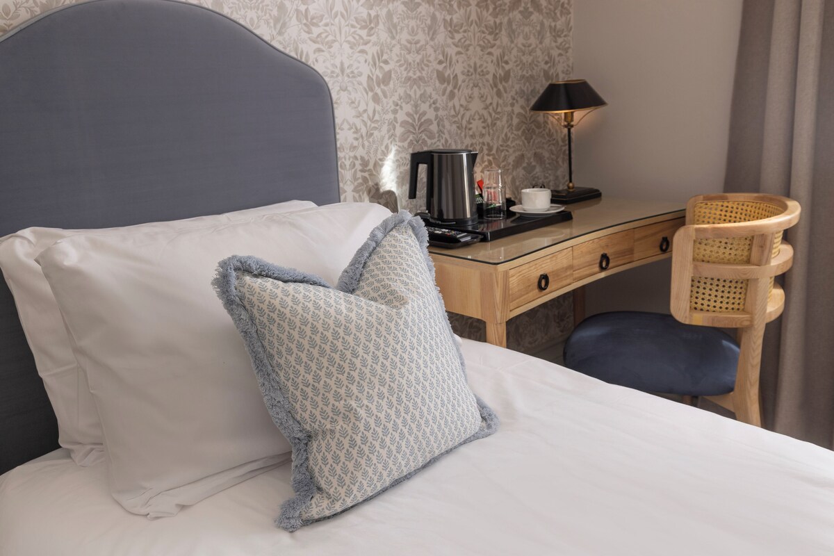 A neatly made bed with pillows, a desk with a lamp, a coffee maker, and a wicker chair.