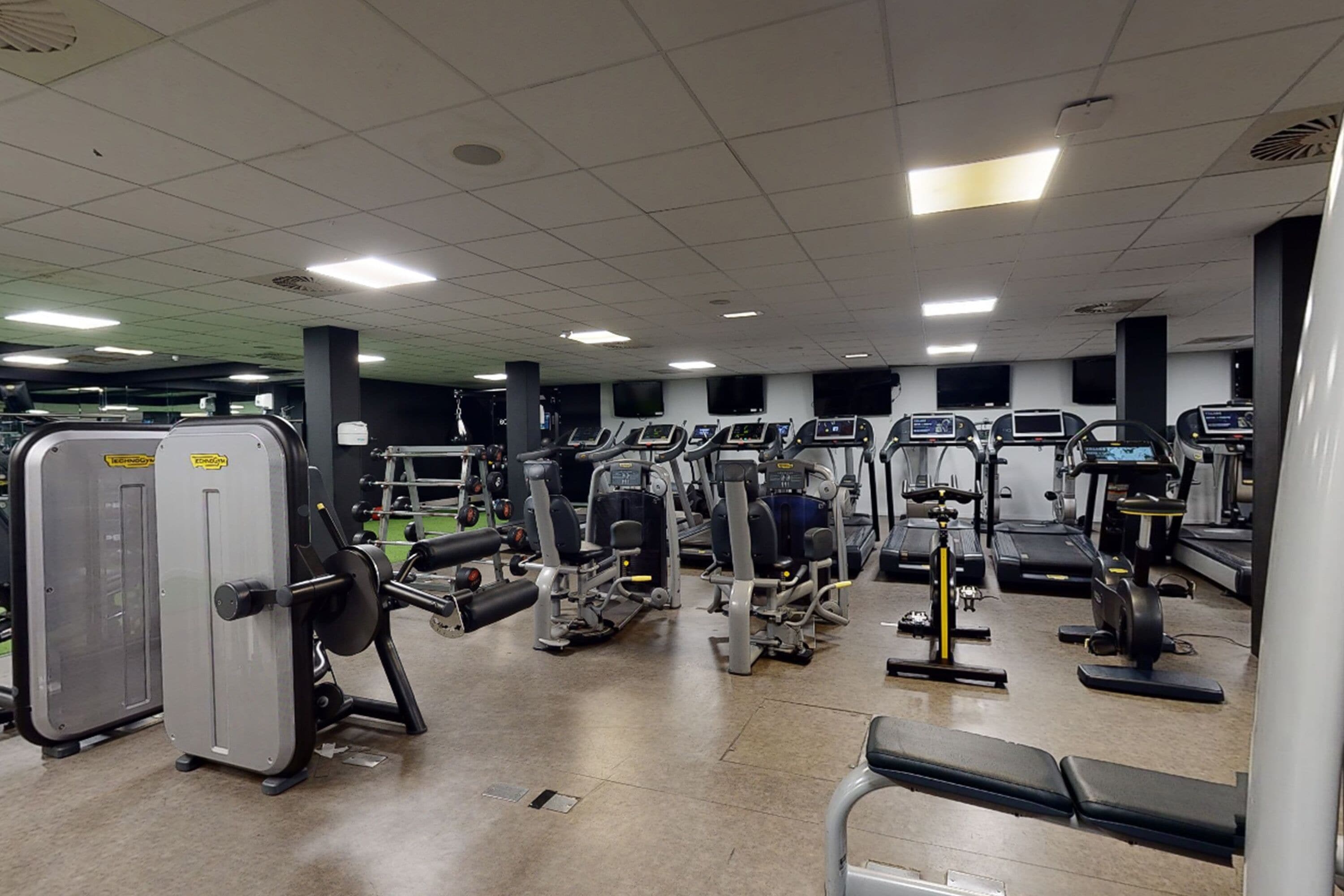 Ein Fitnessstudio mit verschiedenen Trainingsgeräten, darunter Laufbänder, Kraftgeräte und Kurzhanteln.