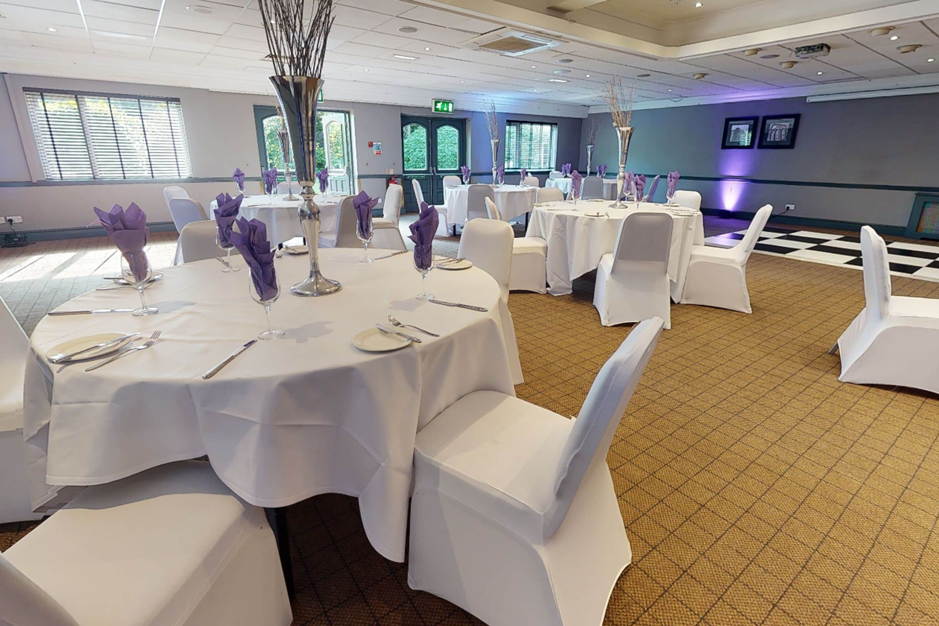 Une salle de banquet avec des tables rondes dressées pour le repas, des chaises blanches et des porte-serviettes violets.