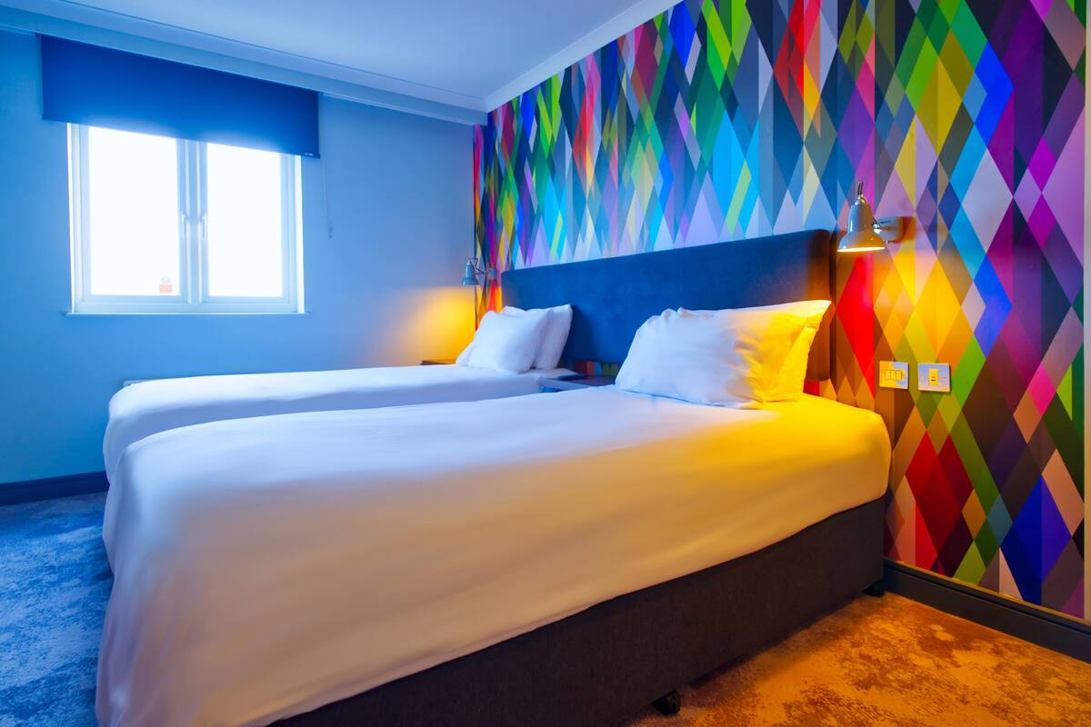 Une chambre d’hôtel avec deux lits, un mur aux motifs géométriques colorés, une fenêtre avec des stores et une lampe sur la table de chevet.