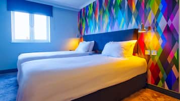 A hotel room with two beds, a colorful diamond-patterned wall, a window with blinds, and a lamp on the nightstand.