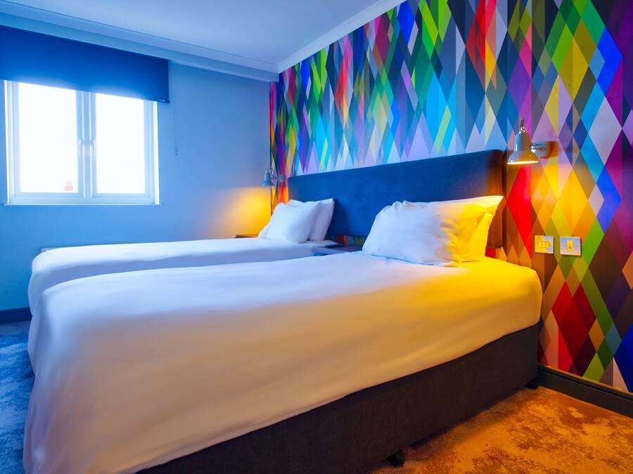 A hotel room with two beds, a colorful diamond-patterned wall, a window with blinds, and a lamp on the nightstand.