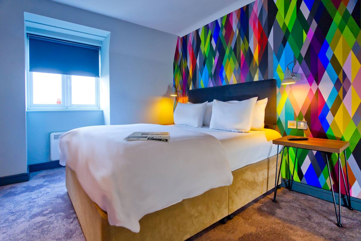 Une chambre d’hôtel avec un lit, une table de chevet, une lampe et un mur aux couleurs vives.
