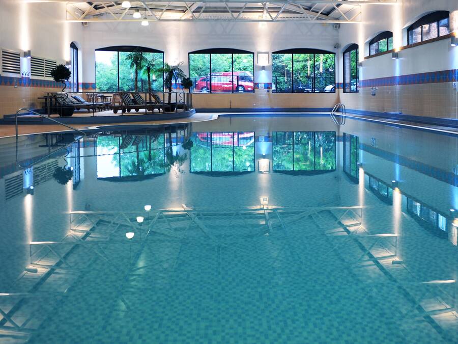 A large indoor swimming pool with clear blue water, sun loungers, and large windows overlooking a red car.