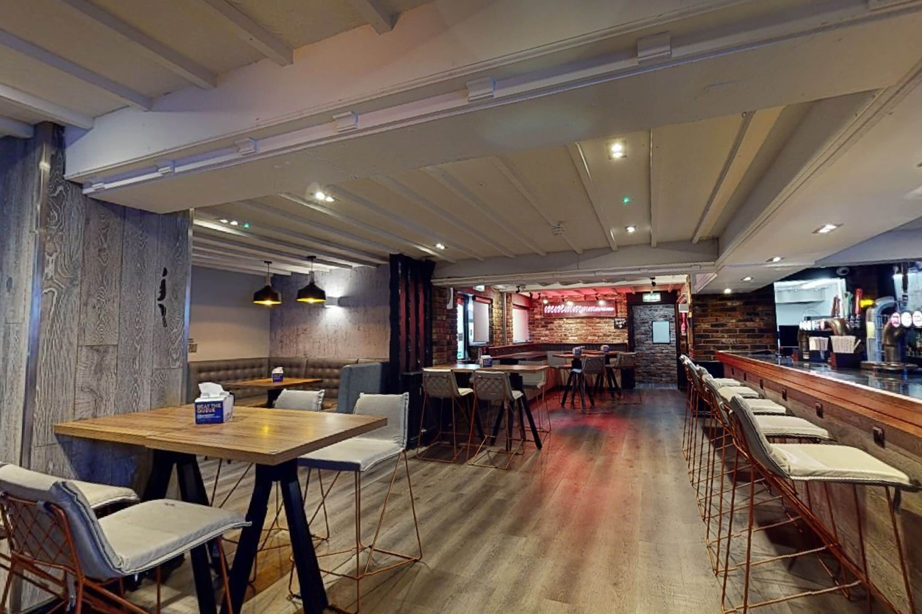 A restaurant with wooden tables and chairs, a bar area, and a brick wall in the background.