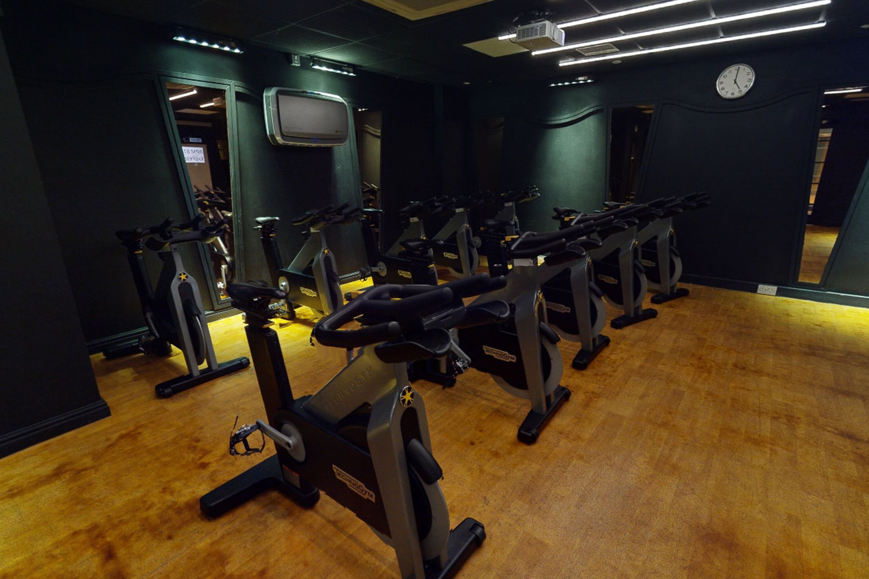 Une salle de sport avec plusieurs vélos d’appartement disposés en rangées, une horloge au mur et un plancher en bois.