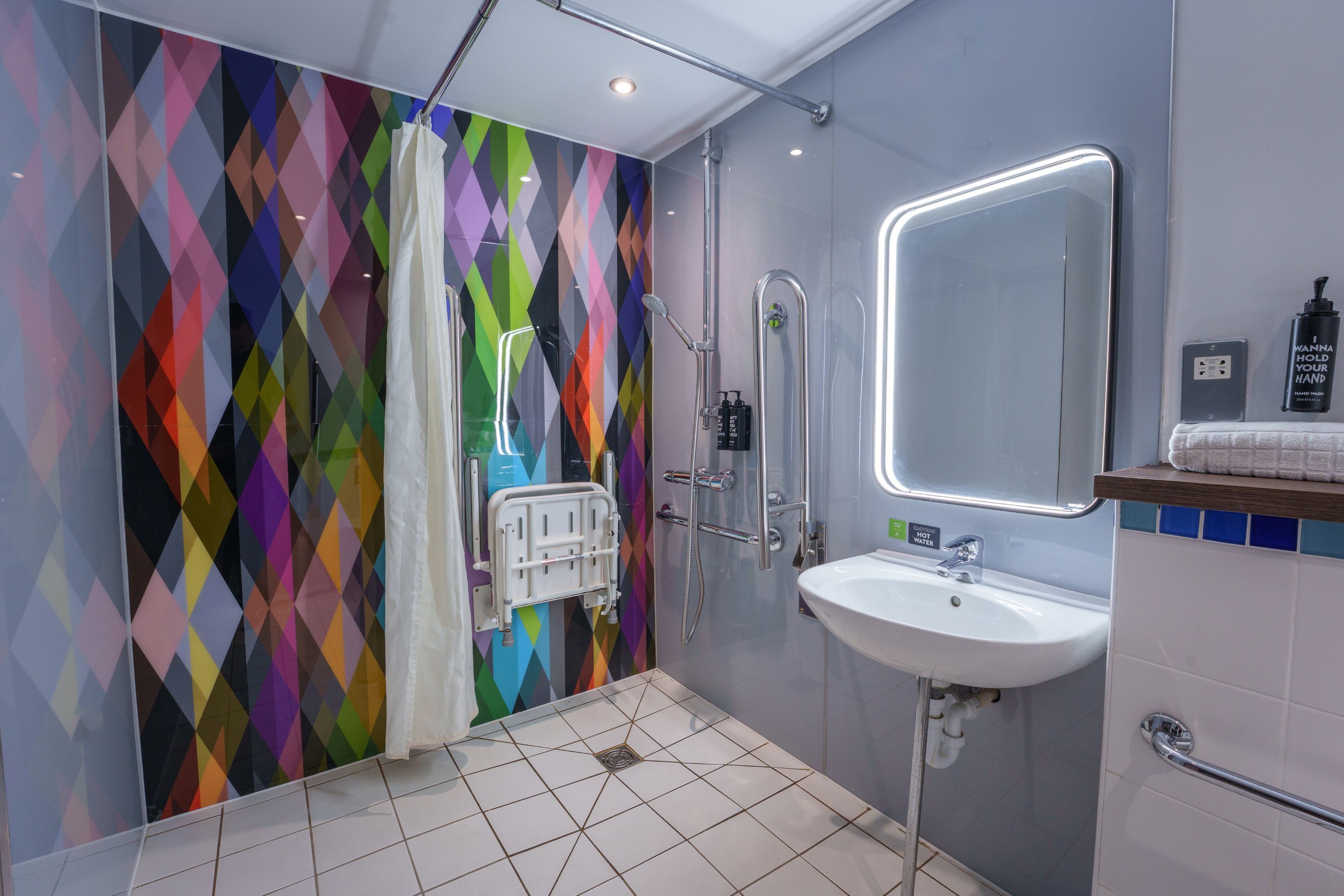 Un bagno moderno con una parete a mosaico colorato a forma di rombo, una doccia, un lavandino con specchio e un dispenser di gel disinfettante.