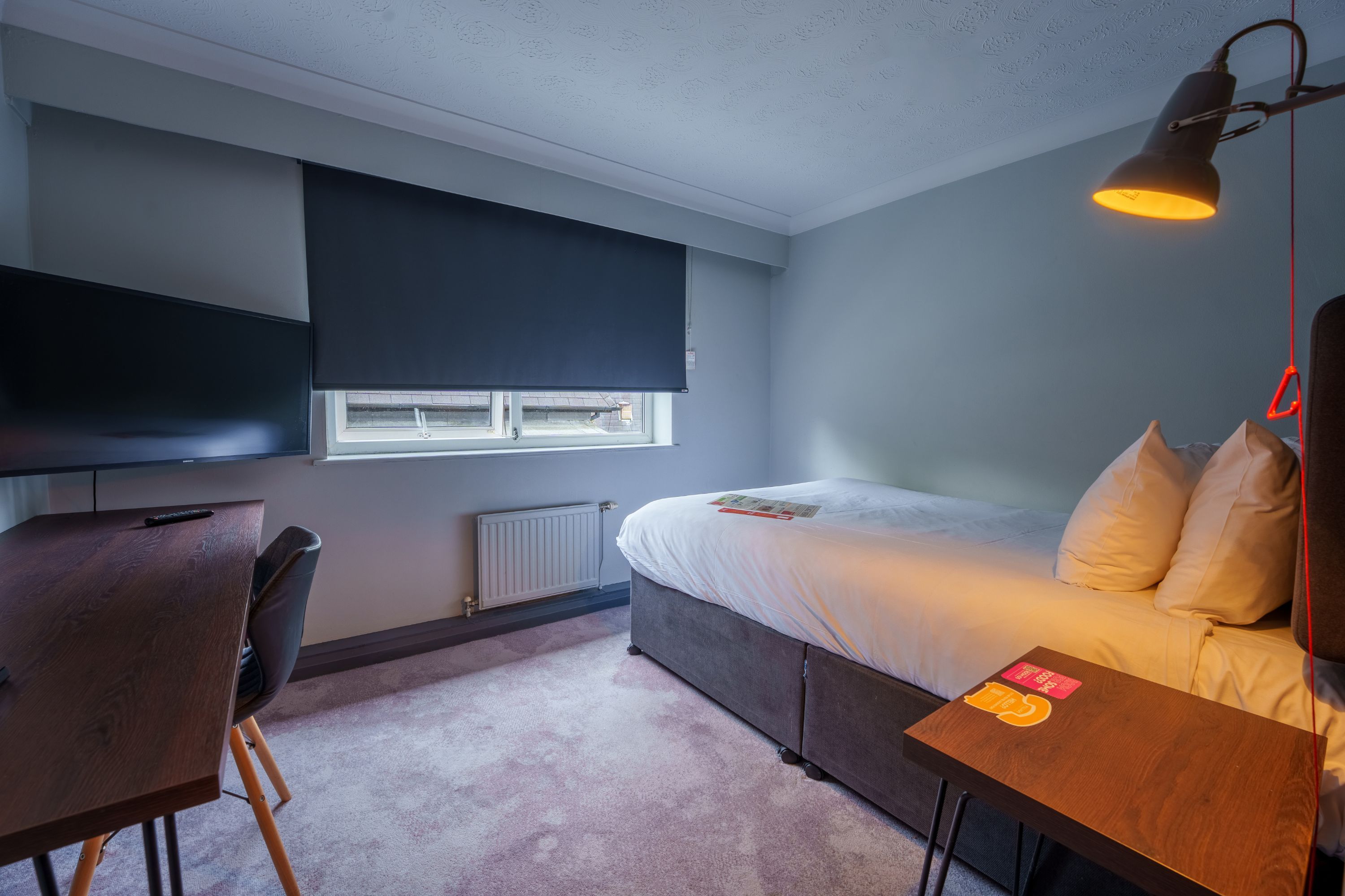 A bedroom with a bed, a desk, a chair, a television, and a window.