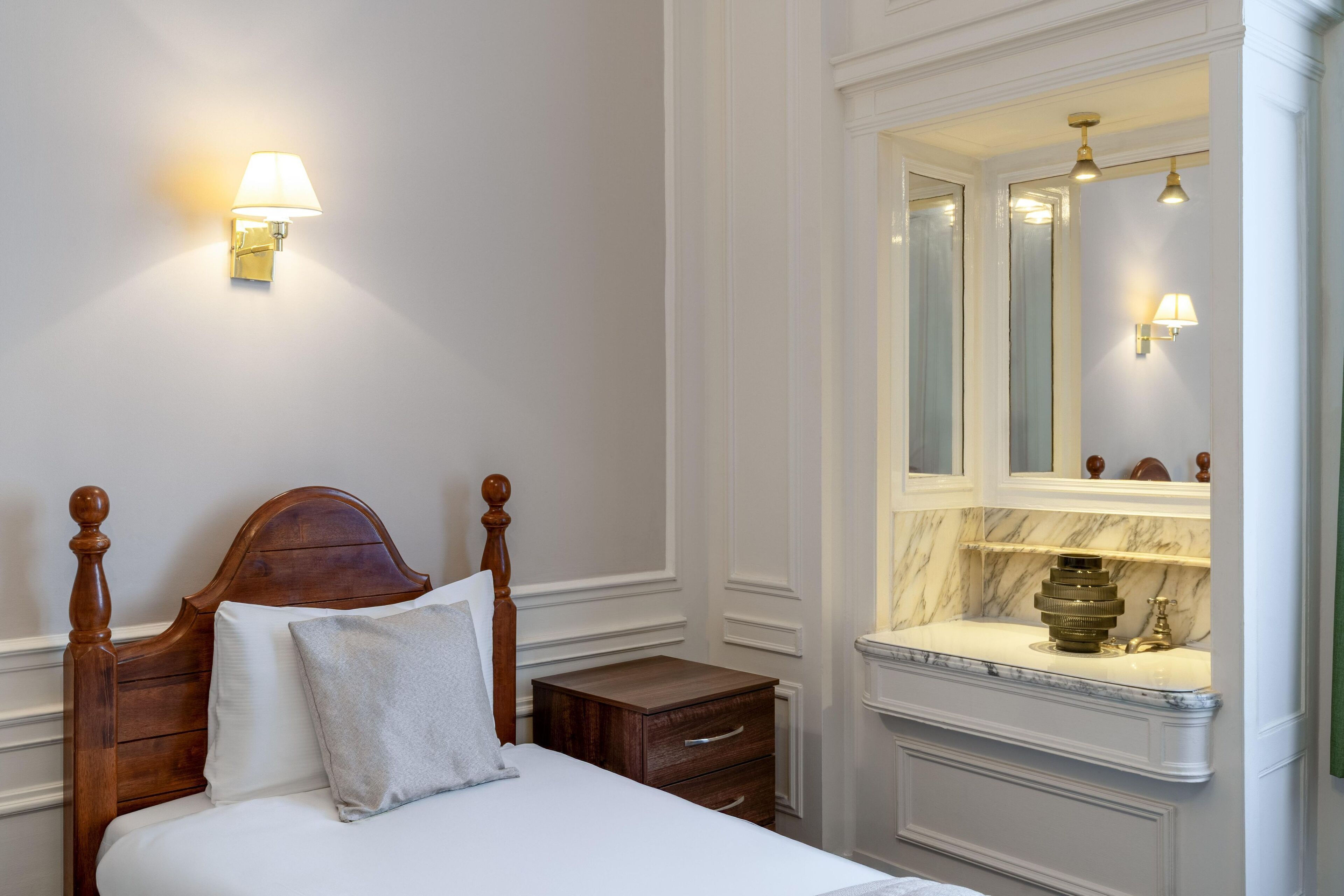 A neatly made bed with a wooden headboard, a nightstand with a drawer, a marble bathroom vanity with a mirror, and wall sconces.