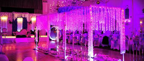 Salón de recepciones de boda con escenario, arreglos florales y candelabro.
