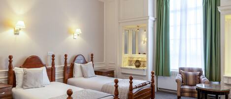 A hotel room with two wooden beds, a sitting area with a chair and table, a window with green curtains, and wall-mounted lamps.