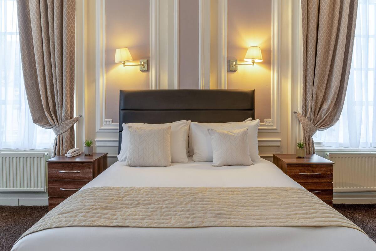 Habitación de hotel con una cama grande, dos mesitas de noche, una lámpara y una ventana con cortinas.