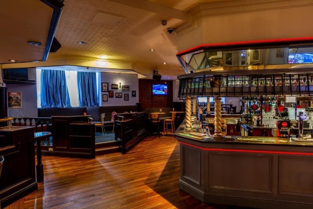 A bar area with a wooden floor, a counter with various bottles, and a television screen.