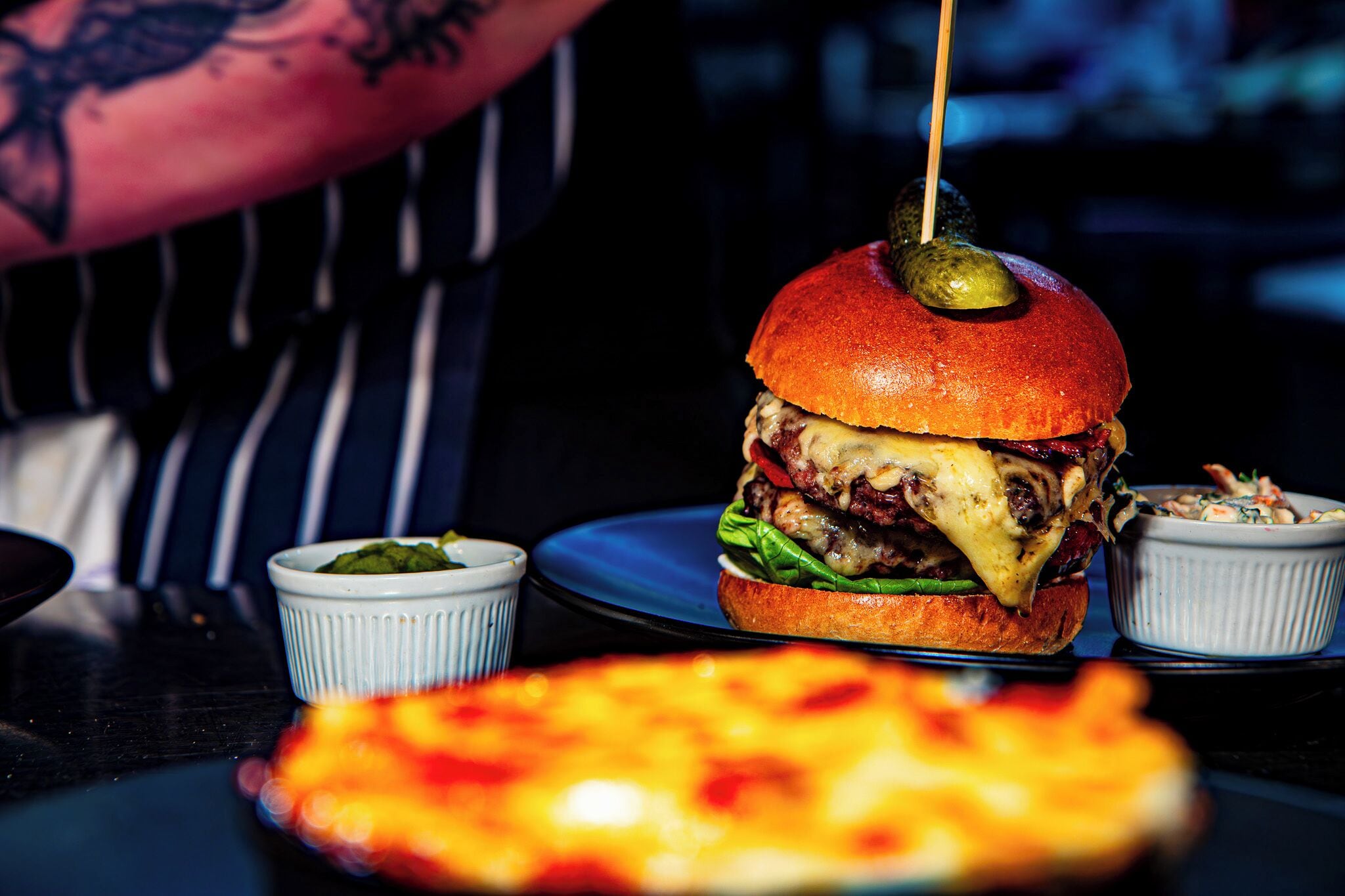 A burger with a toothpick, a small bowl of sauce, and a pizza on a table.