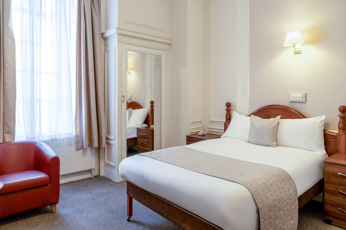 Habitación de hotel con una cama grande, una mesita de noche de madera, un sillón rojo y una ventana con cortinas translúcidas.